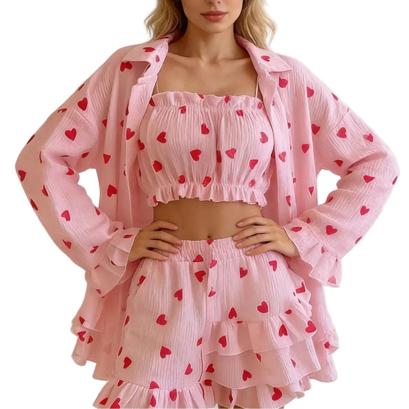 A woman modeling a light pink three-piece matching set featuring a crinkled texture and a red heart pattern, consisting of a long-sleeve button-up shirt, a cropped square-neck top, and a tiered ruffled skirt, set against a white paneled wall.